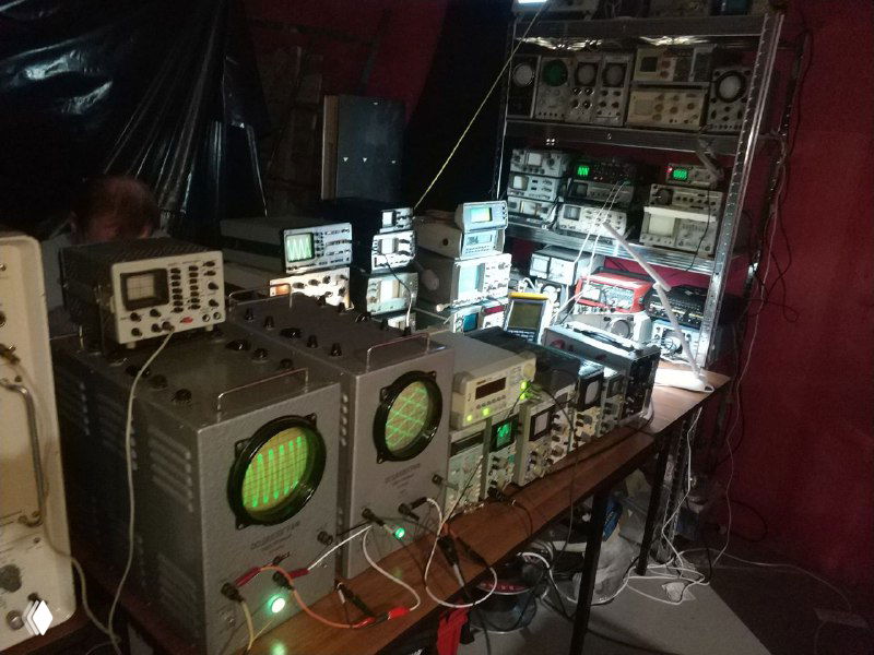 Широкий план: множество осциллографов и мониторов на стеллаже, подсвеченные экраны разной формы и размеров, общий вид экспозиции лабораторного оборудования.