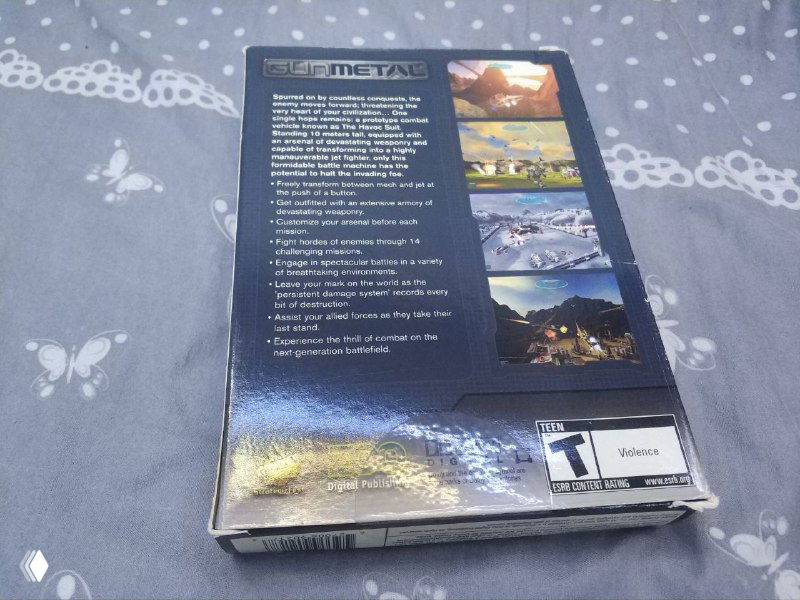 Обратная сторона той же коробки Gun Metal с текстом описания и скриншотами, запечатанная, кадр сделан на столе с узорчатой тканью.