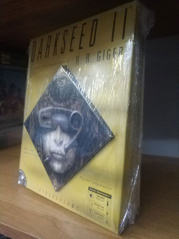 Слегка сбоку: коробка Dark Seed II (MacOS) в блике света на полке; видно оформление лицевой части и отражения плёнки.