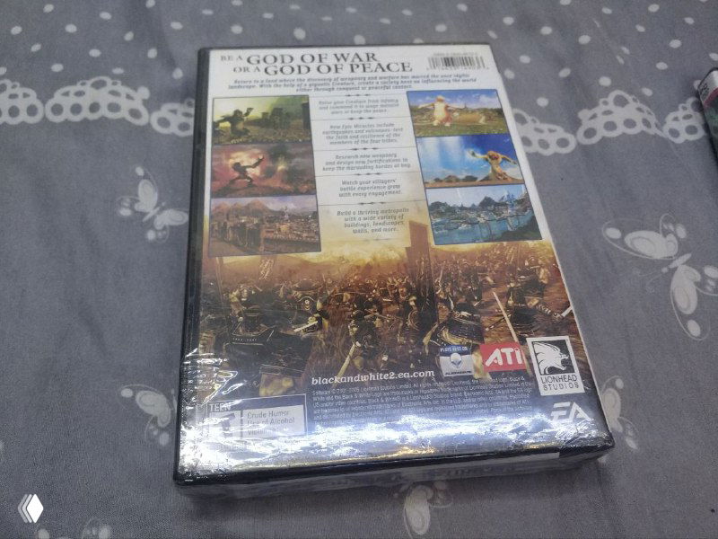 Задняя сторона той же коробки с информацией о содержимом и скриншотами игры, запечатанная в плёнку и положена на серую ткань с узором.