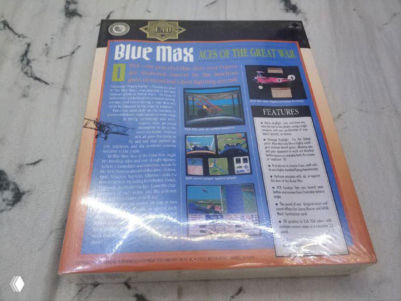 Задняя сторона коробочного издания «Blue Max» в плёнке: текст описания, мини‑скриншоты игрового процесса и блок с характеристиками на фоне столешницы.