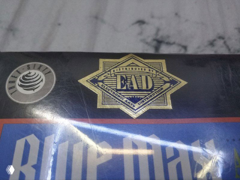 Крупный план верхней части коробки: золотой декоративный знак и печать с надписью, похожей на ранний логотип Electronic Arts Distribution на фоне надписи Blue Max.