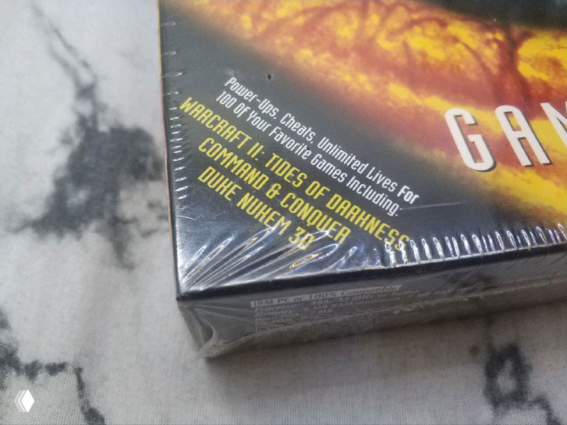 Крупный план угла коробки Game Guru в стрейч‑пленке с видимым печатным текстом о функциях и обещаниями чит‑возможностей; английские фразы частично читаемы.