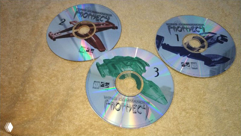 Три компакт-диска Wing Commander Prophecy, пронумерованные и разложенные на фоне ковра: видны декоративные принты и следы использования.
