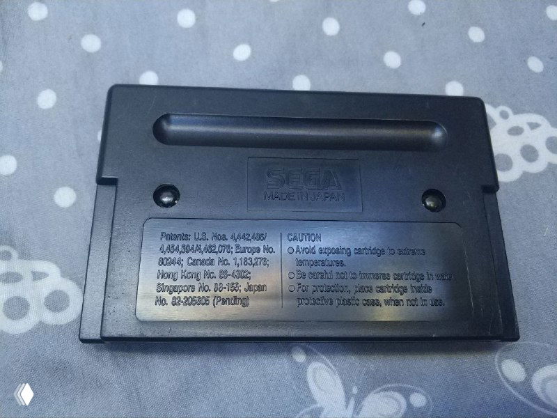 Фото задней стороны картриджа Sega Genesis: пластиковая крышка с наклейкой сервисной информации, видны винты и конструктивные детали корпуса на фоне ткани.
