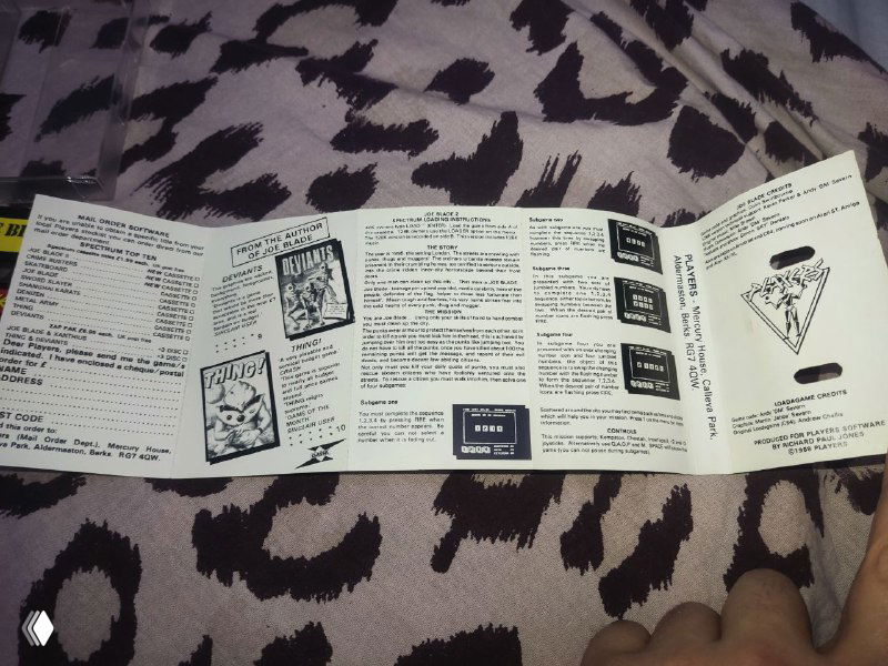 Развёрнутый буклет игры Joe Blade 2: чёрно‑белые инструкции, рекламные блоки и списки, видна рука держит буклет; документ эпохи кассетных релизов.