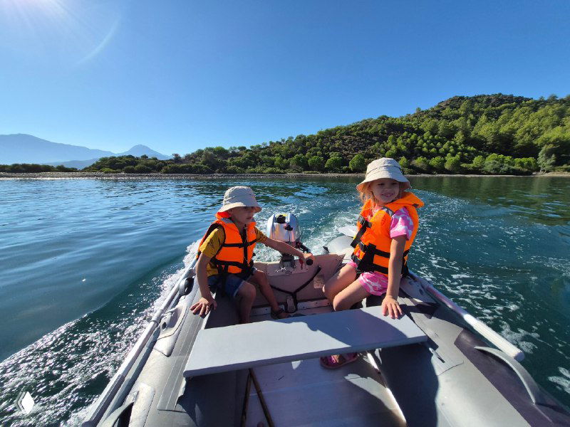 Две ребёнка в ярких спасательных жилетах в небольшой моторной лодке у береговой линии с зелёной растительностью, спокойная вода и яркое дневное солнце, семейный отдых.