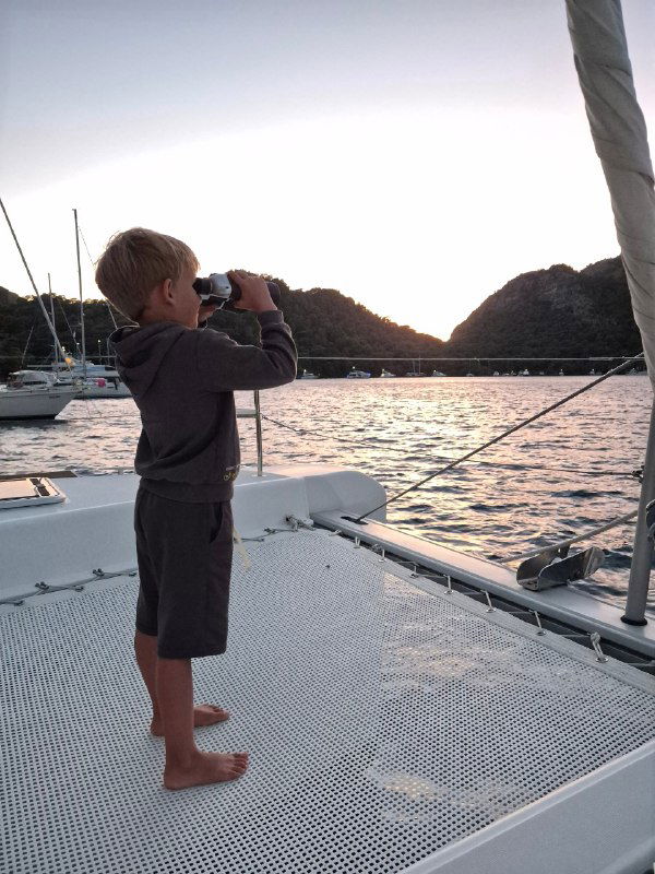 Мальчик в тёплой одежде смотрит в бинокль с носовой части судна на закате, вдали холмы и уходящее солнце, кадр наблюдения и детского любопытства в плавании.