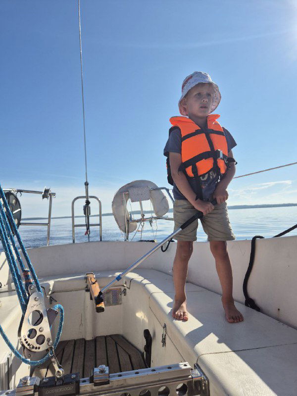 Мальчик в спасжилете стоит у поручней на корме яхты, солнце, море и лёгкий ветер, кадр демонстрирует уверенность и участие ребёнка в морской прогулке.