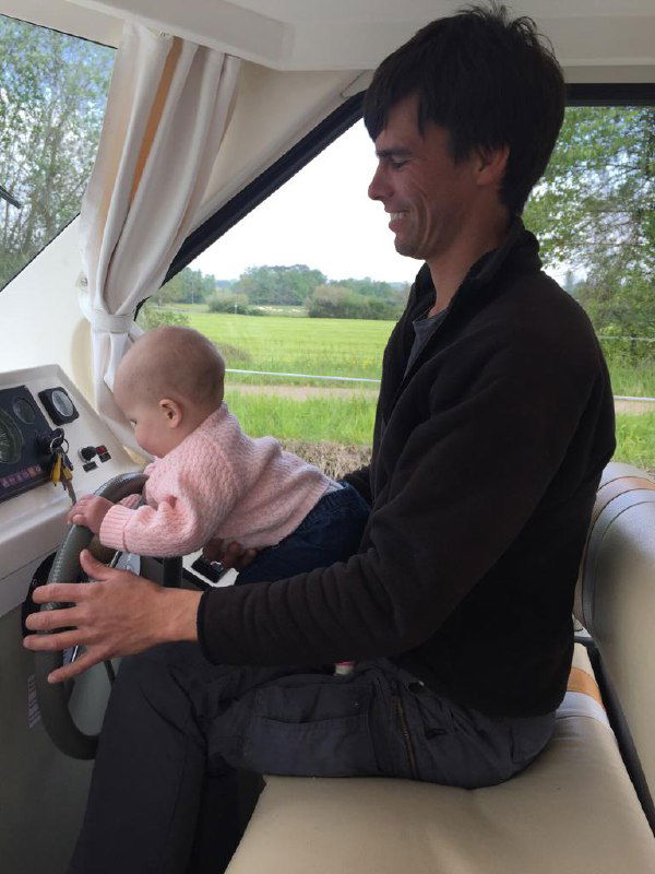 Отец за штурвалом моторной лодки держит на коленях маленькую дочь, внутри видна уютная каюта и окна с видом на зелёный берег, семейная атмосфера на борту.