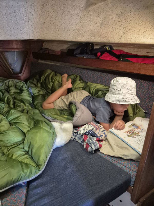 Ребёнок в спальнике в носовой каюте яхты читает книгу, вокруг уютное внутреннее пространство, вещи экипировки и тёплая атмосфера семейного плавания ночью или в тихой стоянке.