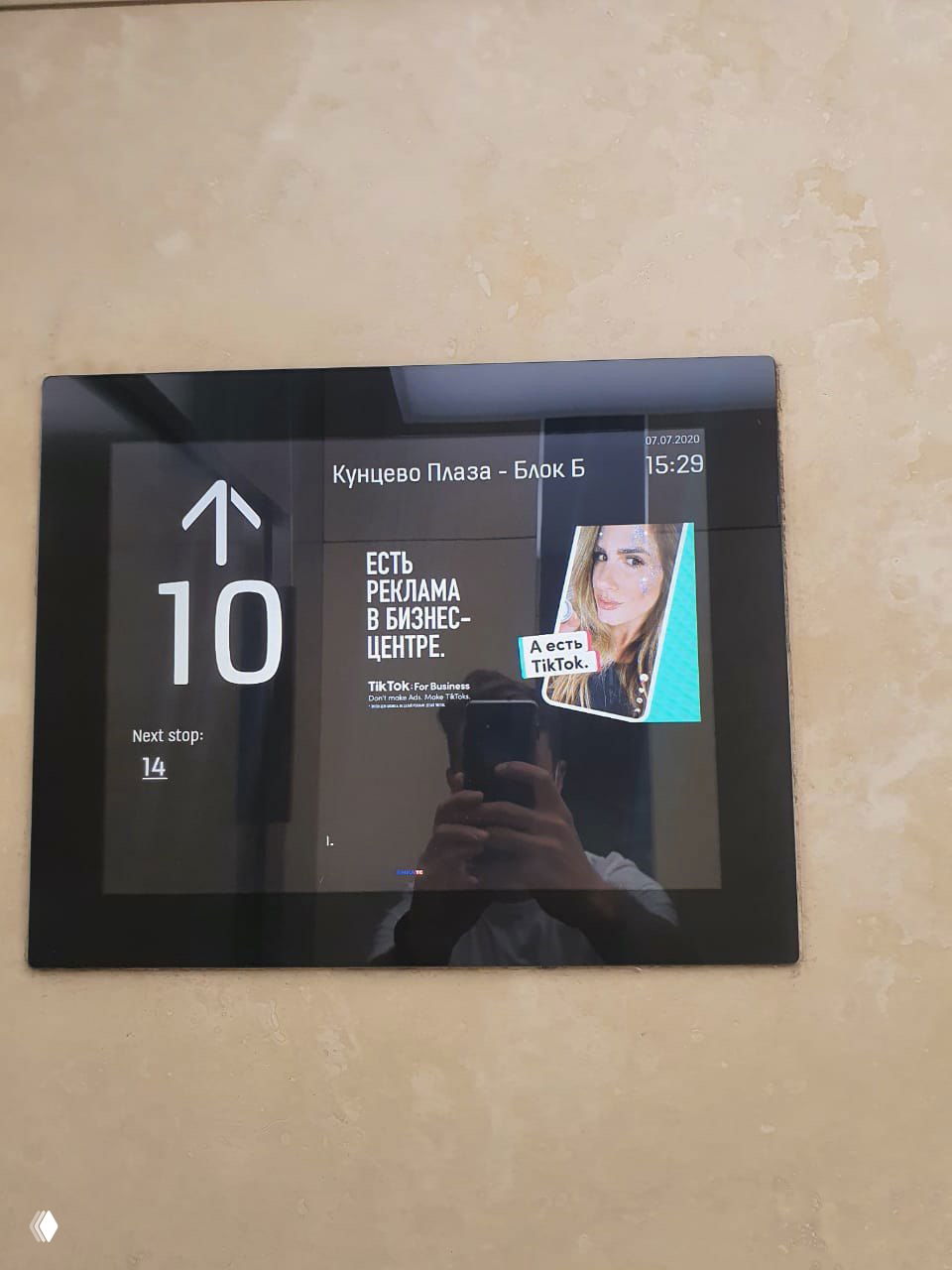 Информационный экран в лифте или холле показывает рекламный слайд TikTok с текстом кампании и маленьким превью ролика с девушкой.