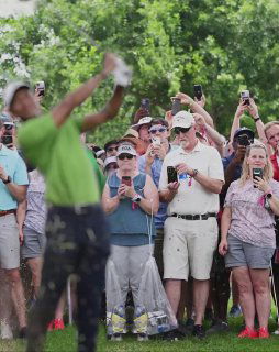 Передний план с мужчиной в зелёной рубашке, за ним плотная толпа болельщиков на гольф-турнире, множество людей держат телефоны и снимают.