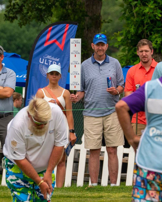 Мужчина в кепке держит банку пива и табличку со спонсорскими логотипами на поле для гольфа; вокруг — другие игроки и зрители.