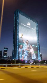 Ночной кадр фасада небоскрёба с огромным билбордом: 3D-изображение Месси с трофеем и логотипом бренда, снято с дороги при уличном освещении.