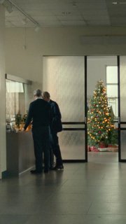Вертикальный кадр из ролика: двое людей у стойки регистрации, в глубине видна украшенная рождественская ёлка в фойе, создаётся бытовая сценка с теплой атмосферой.