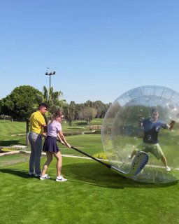 Игроки на поле: женщина готовится ударить по мячу перед прозрачной надувной сферой, в которой стоит человек; солнечный день и зелёное поле.