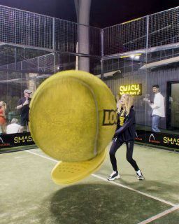 Ночной кадр паддл-корта: крупный желтый теннисный мяч крупным планом и играющие люди на фоне ограждений и рекламных баннеров.