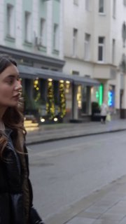 Превью вертикального ролика: молодая женщина в тёмной куртке идёт по городской улице с витринами — кадр для сравнения Reels.