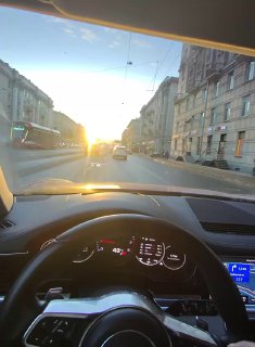 Вид с места водителя: рулевое колесо и панель приборов на фоне городской улицы и заходящего солнца, кадр POV за рулём.