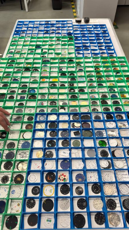 Вид сверху на стол с множеством циферблатов завода «Луч»: десятки лотков зелёного и синего цвета, каждый циферблат с уникальным принтом и оформлением.