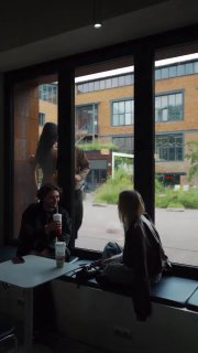 Несколько посетителей кофейни у большого окна; снаружи видно прохожих и современное здание — городской фон для сцены из Reels или сторис.