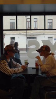 Двое молодых людей в бейсболках пьют напитки за столиком; вид через витрину на городскую улицу создаёт контекст для короткого ролика.