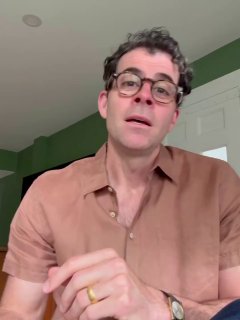 Вертикальный видеокадр: человек в коричневой рубашке и очках говорит в камеру на фоне зелёного интерьера — похоже на отрывок демонстрации перевода в приложении.