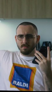 Портретный кадр мужчины в очках и футболке с ярким принтом — герой ролика демонстрирует эмоции при общении с клиентом в офисной обстановке.