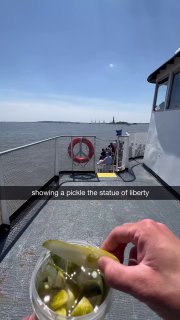 На палубе лодки рука держит банку Grillo's Pickles, вдалеке видна Статуя Свободы и городской горизонт — уличная дегустация огурцов.