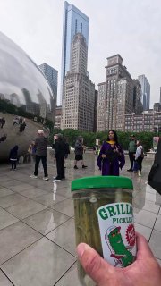 Рука держит банку Grillo's Pickles на фоне популярной городской достопримечательности и небоскрёбов; кадр подчёркивает уличный промо-формат бренда.