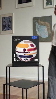 Кадр упаковки La Boule на той же металлической полке, вид с другого ракурса, в кадре видна часть человека и настенные картины; фотография иллюстрирует объект визуального внимания в ленте.