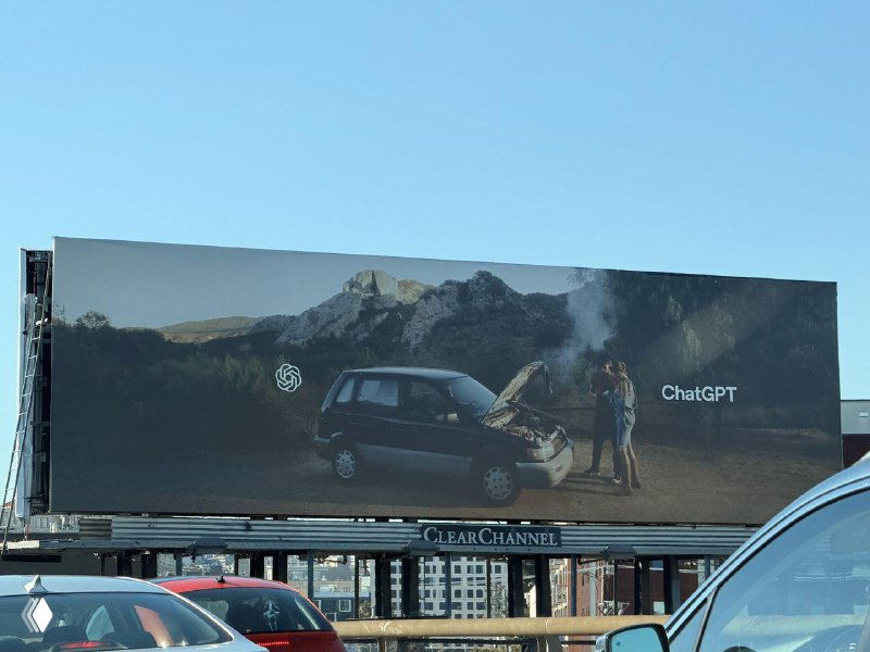 Широкий план билборда с рекламой ChatGPT на фоне города: машина с открытым капотом, пейзаж и минималистичный логотип в правой части панели