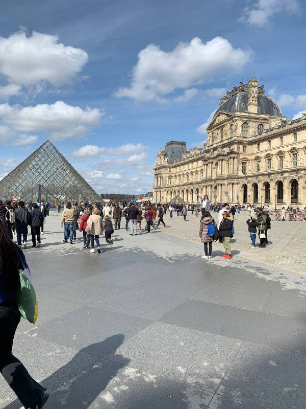 Площадь перед стеклянной пирамидой Лувра: туристы рассредоточены по площади под белыми облаками и солнцем тёплого дня.
