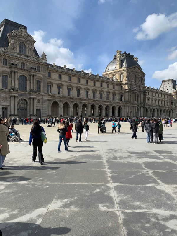 Широкая открытая площадь у центрального крыла Лувра, люди на прогулке и классическая фасадная архитектура на фоне неба.