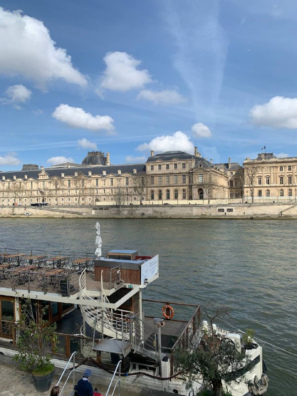 Берег Сены с классическими парижскими домами на противоположной стороне реки, причал и спокойная водная гладь в весенний день.