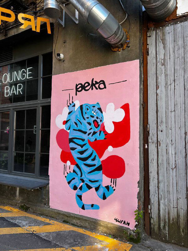 Фасад с настенной росписью Вовы Цыгана: яркая синяя «киса» на розовом фоне у входа в лоунж-бар рядом с уличной сценой в Питере.