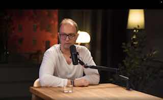 Человек в студии подкаста сидит за деревянным столом перед микрофоном; на столе стакан воды, тёплая интерьерная подсветка, кадр в спокойной обстановке.