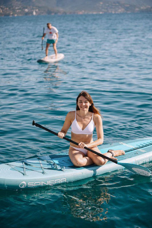 Девушка сидит на SUP-доске с веслом в руках на спокойной поверхности моря, позади виднеется городской берег и другие отдыхающие на воде.