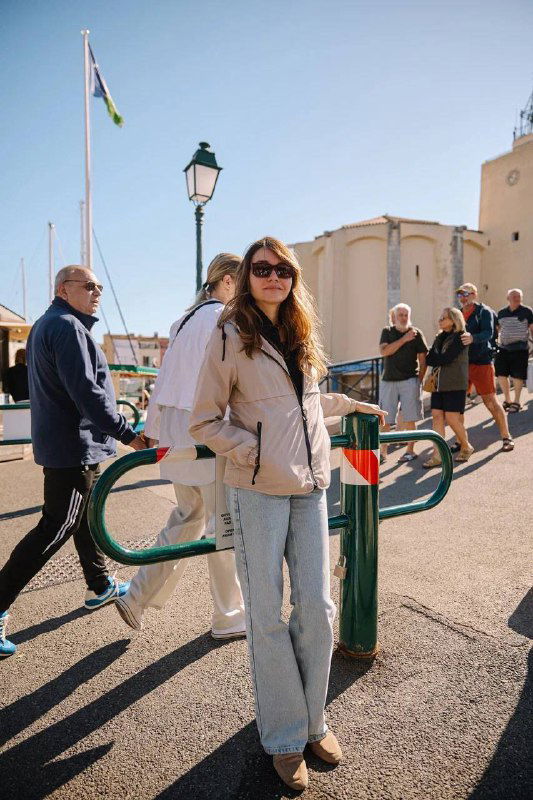 Автор идёт по набережной в лёгкой куртке и джинсах; вокруг прохожие, уличные фонари и средиземноморская архитектура городка.