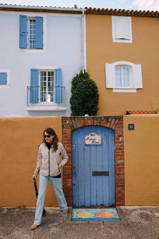Яркий фасад дома с голубой дверью и ставнями; женщина на переднем плане в повседневной одежде рядом с живописной архитектурой.