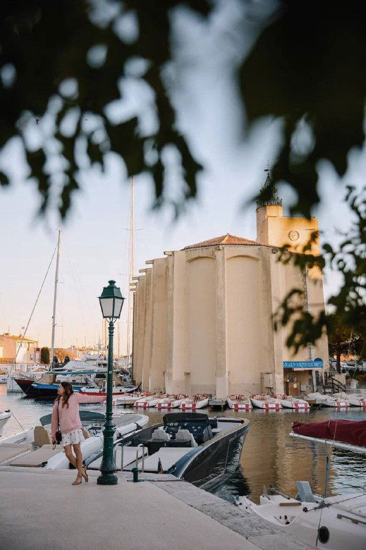 Набережная с причалом и лодками, исторические здания на заднем плане; женщина идёт вдоль причала в тёплом свете вечернего солнца.