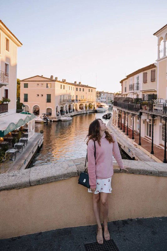 Женщина стоит на небольшом мостике над каналом, по сторонам магазинчики и дома; мягкий дневной свет и спокойная туристическая сцена.