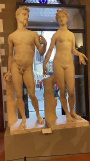 Крупный план мраморных статуй в музее Барджелло во Флоренции: пара античных фигур на пьедестале в музейном интерьере с витражом позади.