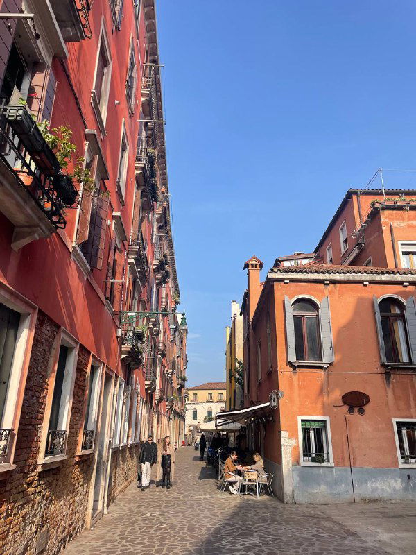 Узкая венецианская улочка с красными фасадами, балконами и маленькими кафе; прохожие идут по брусчатке под чистым голубым небом.