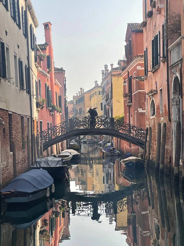 Небольшой каменный мост через тихий канал, спокойная зеркальная вода и отражения тёплых фасадов старых венецианских зданий.