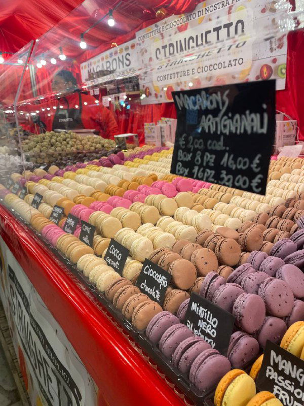 Яркий прилавок с макаронами и десертами на рождественском рынке: разноцветные макарунсы и ценники на витрине палатки в красной ткани.