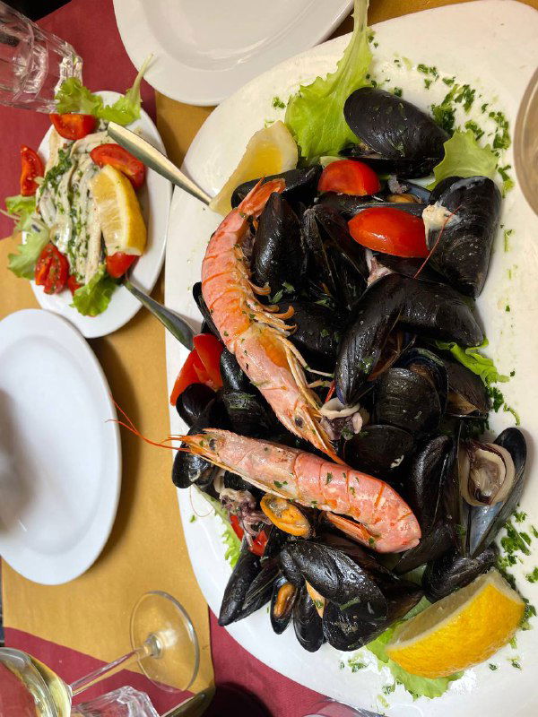 Большая тарелка с мидиями, креветками и лимоном на столе в ресторане Генуи; видна свежая морская подача и салат рядом.