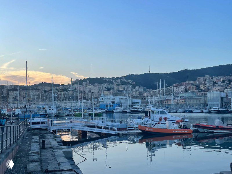 Порт с причаленными яхтами на фоне холмов Генуи в вечернем свете — вид на гавань и морскую линию города.