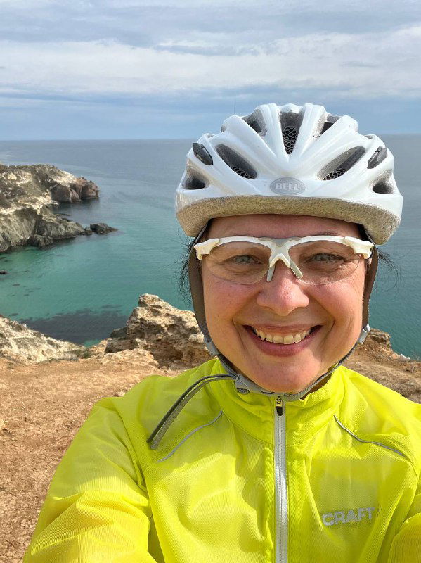 Крупный план улыбающейся женщины в велошлеме и очках на фоне моря и скал, видно салатовую ветровку.